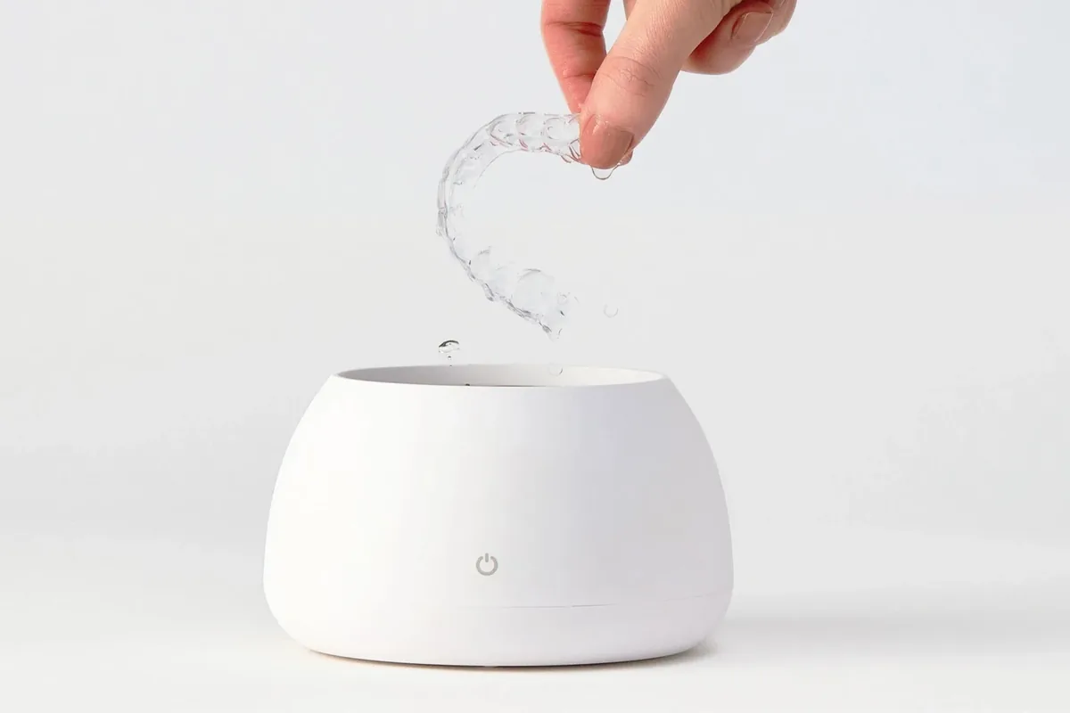 A hand lowering a clear aligner into a white Zima Dental Pod.
